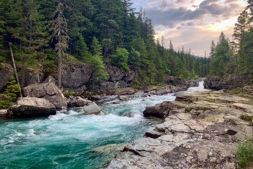 A serene river flows through rocky terrain, surrounded by lush green trees under a cloudy sky.