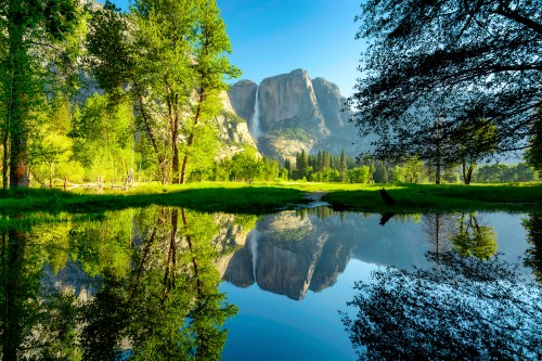 A serene landscape featuring mountains reflected in a calm pond, surrounded by lush greenery and clear blue skies.