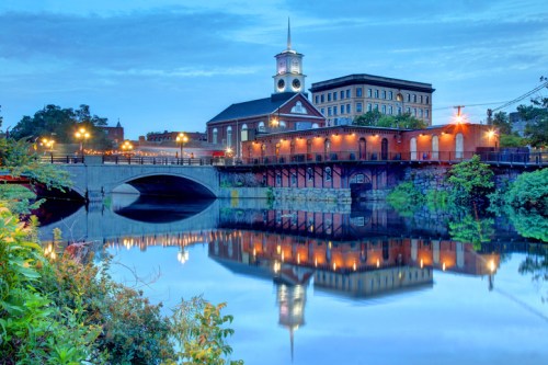 A serene river scene at dusk, featuring a historic building and bridge, with reflections in the calm water.