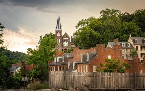 A scenic view of brick buildings with a church steeple in the background, surrounded by lush greenery and a cloudy sky.