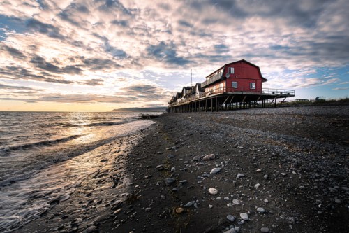 A red house on stilts by the shore, with a rocky beach and a cloudy sky reflecting the sunset over the water.