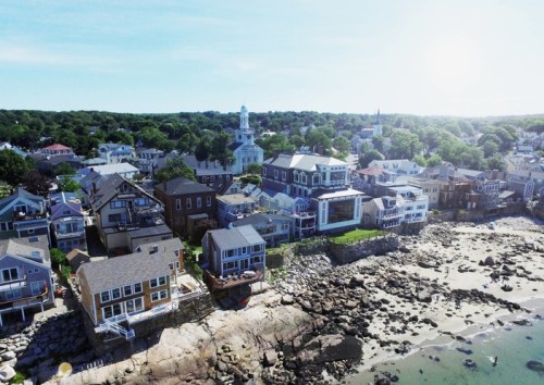 Aerial view of a coastal town with houses, rocky shoreline, and a church steeple under a bright sky.