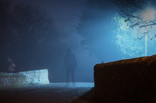 A shadowy figure stands on a foggy bridge, illuminated by a blue streetlight, creating an eerie atmosphere.
