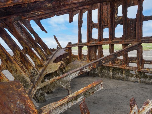 Rusty shipwreck interior with exposed beams and weathered textures, set against a sandy beach and cloudy sky.