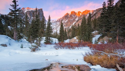 Snow-covered landscape with mountains and pine trees, illuminated by a colorful sunrise. A stream flows through the scene.