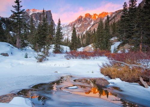 Snow-covered landscape with a stream reflecting mountains and colorful sky at sunrise, surrounded by evergreen trees.
