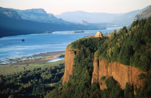 A scenic view of a river valley surrounded by mountains, with a historic structure perched on a cliff.