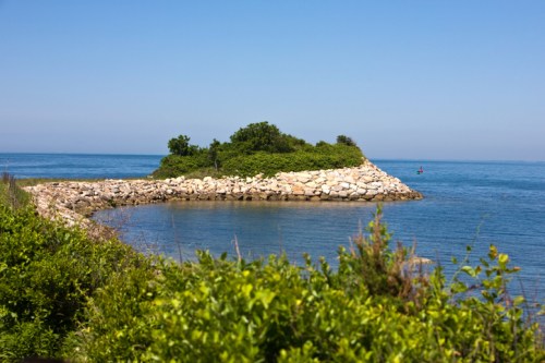 A rocky shoreline extends into calm blue waters, surrounded by lush greenery under a clear blue sky.