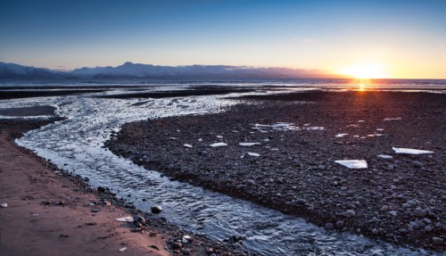 A serene beach scene at sunset, with a winding stream and rocky shoreline under a colorful sky.