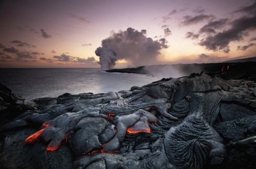 Lava flows on rocky shore at sunset, with smoke and clouds rising from a volcano in the background.
