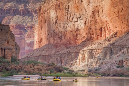 Rafts float on a river surrounded by towering red rock cliffs and lush greenery in a canyon landscape.