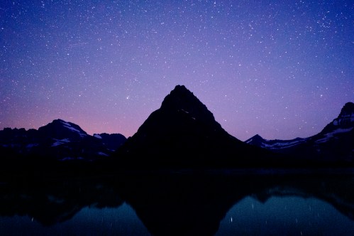 A serene mountain silhouette against a starry night sky, reflecting in a calm lake below.
