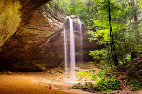 A serene waterfall cascades from a rocky overhang into a lush, green forest setting.