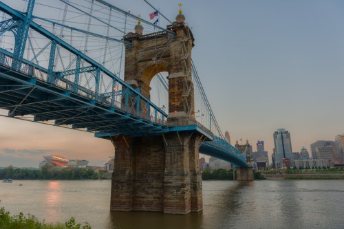A blue suspension bridge spans a river, with a city skyline in the background during sunset.