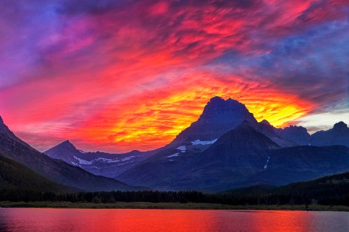 Vibrant sunset over mountains, reflecting in a calm lake, with colorful clouds illuminating the sky.