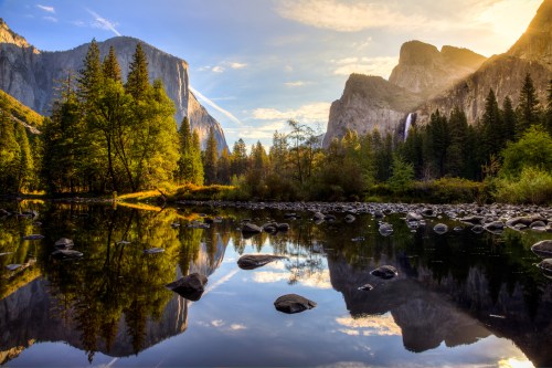 A serene landscape of mountains and trees reflecting in a calm river at sunset, with a waterfall in the background.