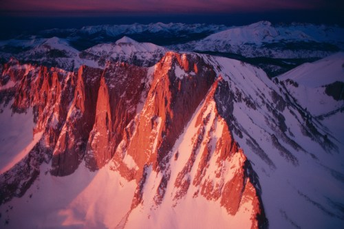 Snow-covered mountain peaks bathed in warm sunset light, with a dramatic landscape of ridges and valleys.