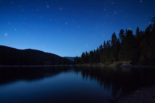 A serene lake at dusk, reflecting stars and trees under a deep blue sky.