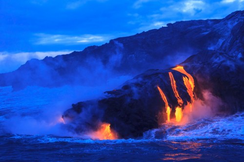 Lava flows into the ocean, creating steam and glowing orange against a dark rocky landscape under a blue sky.