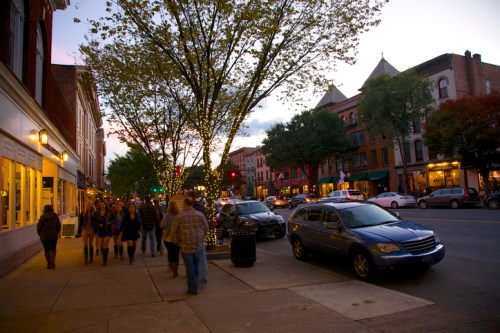 A bustling street scene at dusk, with people walking, trees lining the sidewalk, and shops illuminated.