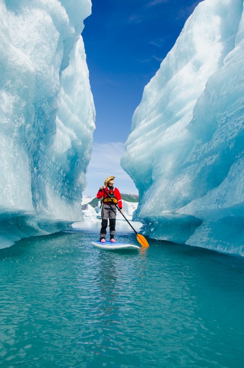 A person paddleboarding between towering ice formations in a serene glacial landscape under a clear blue sky.