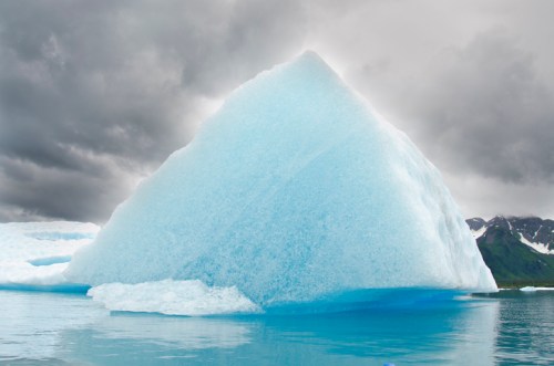 A large, pyramid-shaped iceberg floats in calm waters under a cloudy sky, surrounded by mountains.