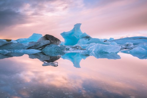 A serene landscape featuring icebergs reflected in calm water under a colorful sky at sunset.