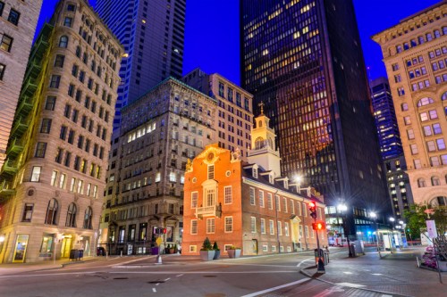 Historic building illuminated at dusk, surrounded by modern skyscrapers in a bustling city street.