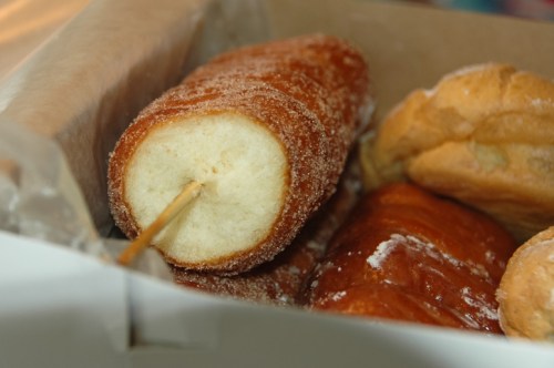 A close-up of a sugary pastry on a stick, surrounded by other baked goods in a box.