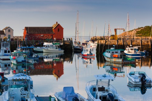 A peaceful harbor scene with boats and a red building reflecting in calm water under a clear sky.