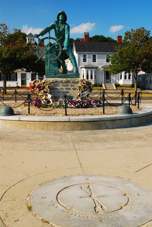 A bronze statue of a sailor at a ship's wheel, surrounded by flowers and a compass rose on the ground.