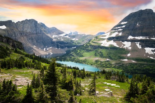 A scenic mountain landscape with a lake, lush greenery, and a colorful sunset sky. Snow-capped peaks in the background.