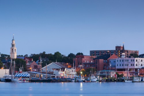 A scenic waterfront view at dusk, featuring boats, buildings, and a clock tower against a clear blue sky.