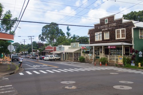 A small town street with shops, a historic building, and parked cars under a clear sky. Pedestrians cross at a crosswalk.
