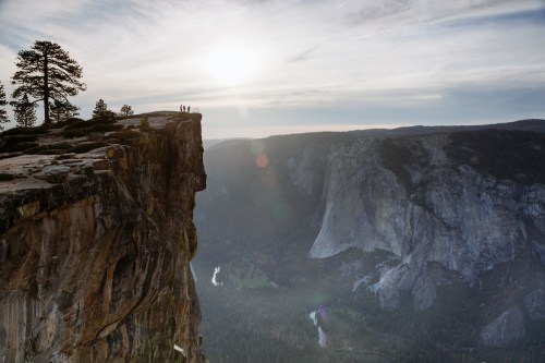 Silhouetted figures stand on a cliff edge at sunset, overlooking a vast valley and distant mountains.