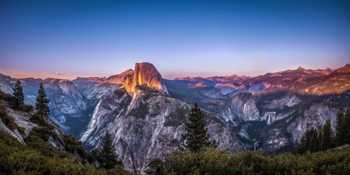 A panoramic view of Yosemite National Park at sunset, featuring Half Dome and surrounding mountains bathed in warm light.