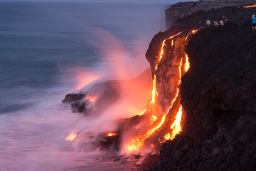 Lava flows down a rocky cliff into the ocean, creating steam and a dramatic contrast against the dark landscape.