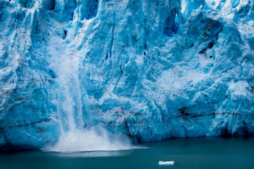 A massive glacier with bright blue ice, cascading water, and a small iceberg in the foreground.