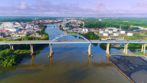 Aerial view of a river with a bridge, industrial buildings, and green landscapes under a cloudy sky.