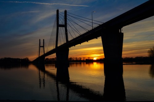Silhouette of a bridge at sunset, reflecting in calm water with colorful sky and clouds.