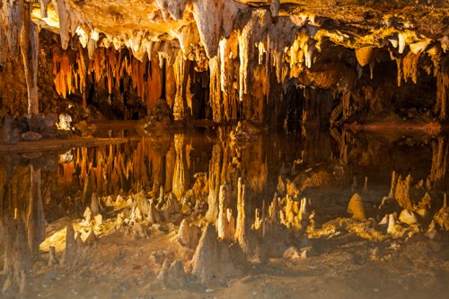 Stalactites and stalagmites reflect in a calm underground pool within a cave, illuminated by warm lighting.