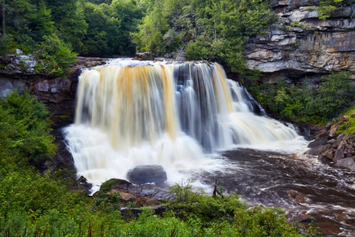 A cascading waterfall flows over rocky cliffs, surrounded by lush green trees and foliage.