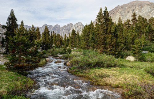 A serene mountain landscape featuring a flowing stream, lush greenery, and towering peaks in the background.