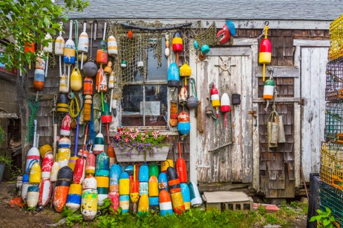 Colorful fishing buoys and nets adorn a rustic wooden shed, with flowers in a window box adding a touch of charm.