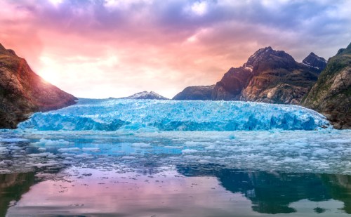 A stunning glacier reflects in calm waters, surrounded by mountains under a colorful sunset sky.