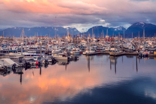 A bustling marina filled with boats, reflecting colorful clouds and mountains in the background at sunset.