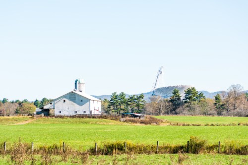 A white barn with a green roof stands in a green field, with mountains and a radio tower in the background.