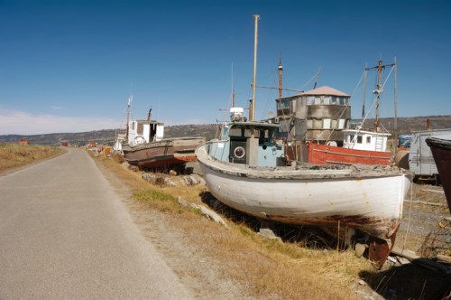 Abandoned boats lined along a dirt road, with a clear blue sky and distant hills in the background.