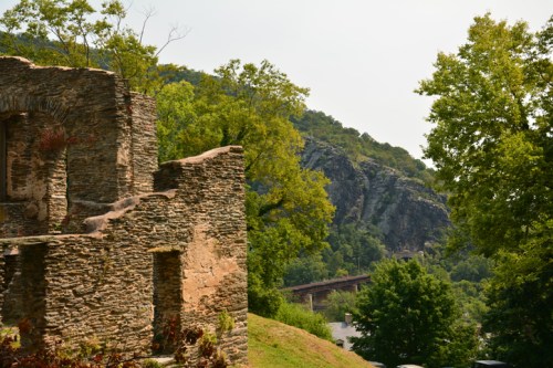 Ruins of a stone structure surrounded by trees, with a rocky hill and bridge in the background.