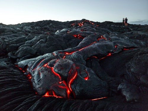 Lava flows across a rocky surface, glowing red in the cracks, with two people observing in the background.